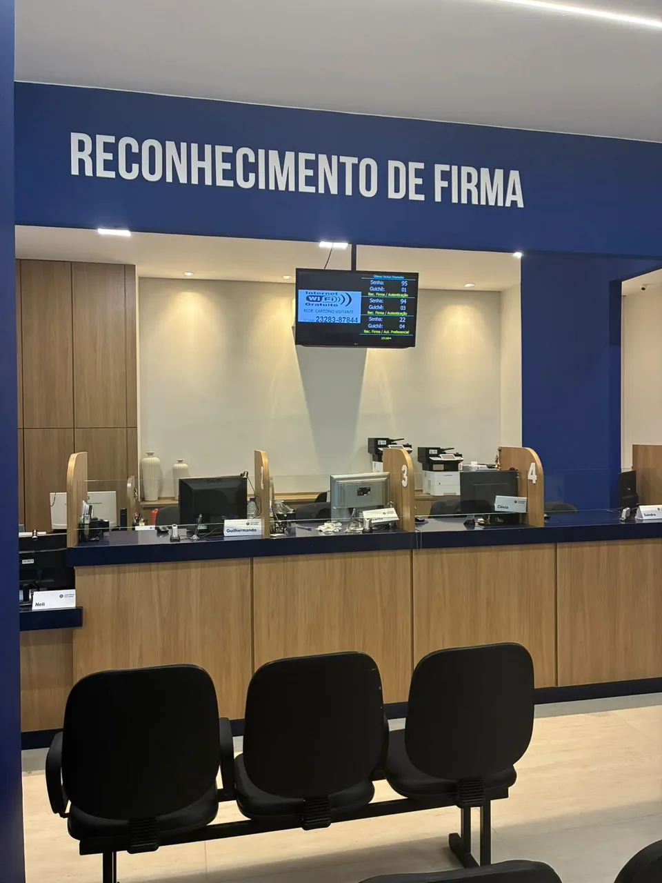 Balcão de atendimento em madeira com painel de senha e cadeiras de espera